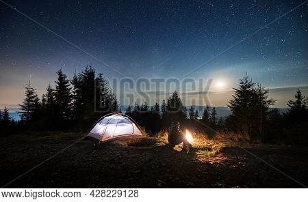 Side View Of Young Man Sitting On Grass Near His Lighted Tent And Watching The Night Stars And Moon 