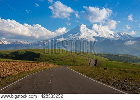 An Asphalt Road Leading To Snow-capped Mountain Peaks. A Summer Day. Elbrus On The Background Of Gre