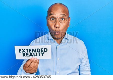 Middle age latin man holding paper with extended reality message scared and amazed with open mouth for surprise, disbelief face 