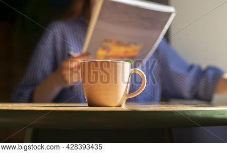 Coffee Cup On Wood Table In Cafe, Blurred Female Hands With Texbook Or Notebook, Reading And Writing