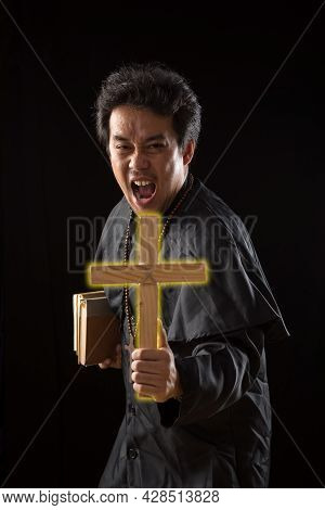 Young Asian Priest Show Lighting Crucifix While Holding Bible To Exorcise Ghost On Black Background