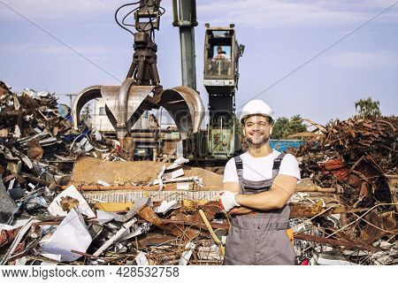 Portrait Of Worker Standing In Metal Junk Yard With Crane Lifting Scrap Metal For Recycling.