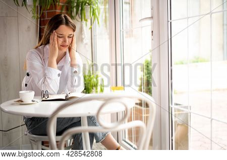 Young Tired Woman In Depression With A Headache Sits In A Modern Cafe Holding Her Head With Her Hand