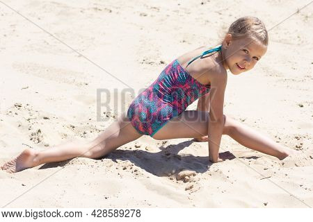 Caucasian Little Girl Of Five Years Old In Swimsuit On Sand Beach Making Leg-split In Summertime