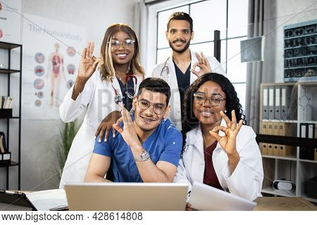 Positive Male And Female Doctors In Medical Clothes Posing At Table With Modern Gadgets, Smiling And