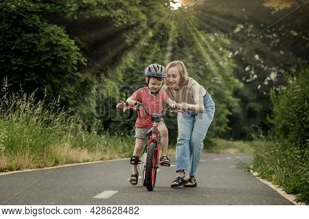 Mother Teaching Son To Ride Bicycle. Happy Cute Boy In Helmet Learn To Riding A Bike On The Bike Pat