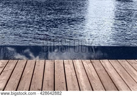The Water Surface Is Dark Blue Next To The Wooden Sidewalk. Birch Of Flat Black Stone