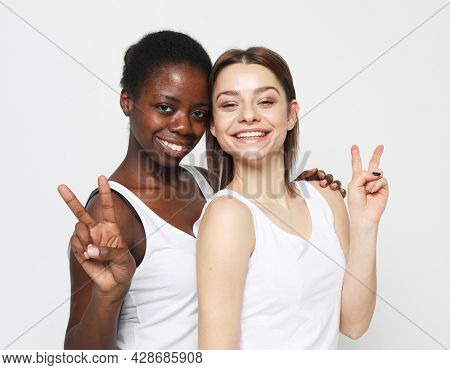 Two young multiracial women wearing white shirts standing together and smiling at camera, show v-sign,
