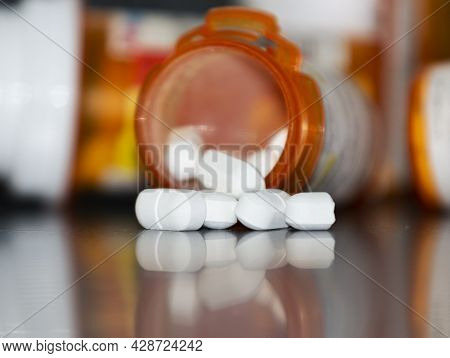 White Drugs Spilling Out Of A Prescription Pill Bottle Lying On Its Side And Reflecting Off Of A Bla