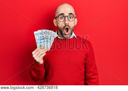 Young bald man holding peruvian sol banknotes scared and amazed with open mouth for surprise, disbelief face 
