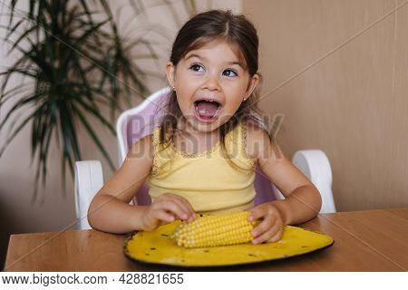 Happy Little Girl Hold Fresh Corn In Hand And Smile. Female Kid Thumbs Up. Summer Mood At Home