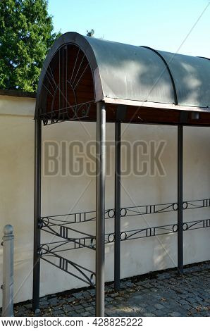 Metal Bus Stop By The Beige Plaster Wall. The Roof Is Made Of Sheet Metal Bent Into An Arch. Cylinde