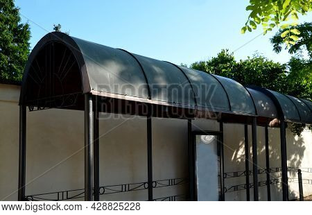 Metal Bus Stop By The Beige Plaster Wall. The Roof Is Made Of Sheet Metal Bent Into An Arch. Cylinde