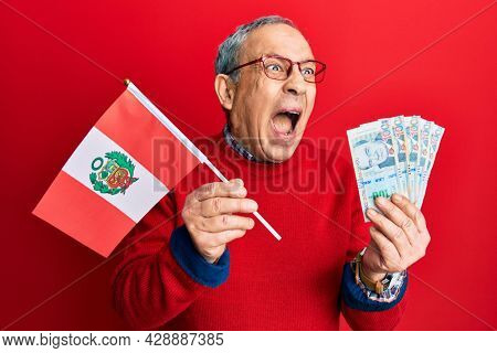 Handsome senior man with grey hair holding peru flag and peruvian sol banknotes angry and mad screaming frustrated and furious, shouting with anger. rage and aggressive concept. 