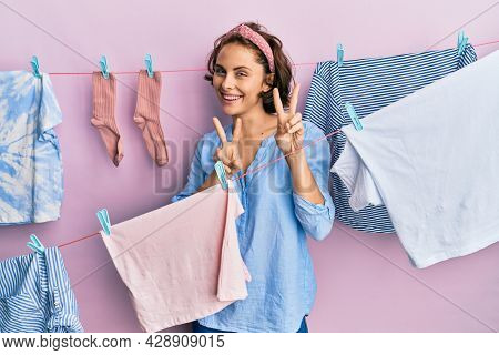 Young brunette woman doing laundry around string hangs smiling looking to the camera showing fingers doing victory sign. number two. 