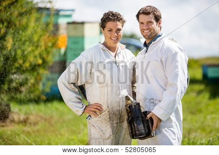 Portrait of happy male and female beekeepers with smoker standing at apiary