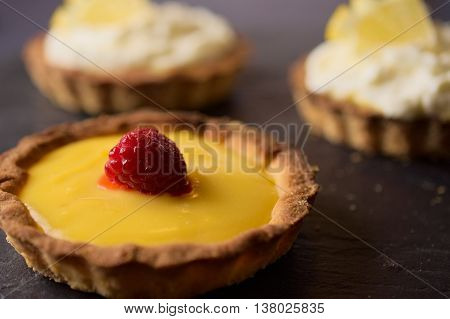 Delicious sweet dessert fresh cheese with lemon and vanilla with raspberry tarts on dark stone slate. Selective focus on lemon slice. Concept of Home baking. Concept of home made desserts.