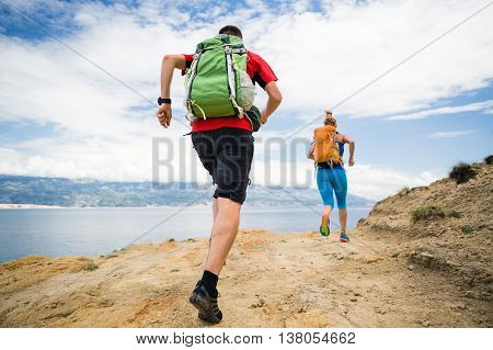Couple runners running with backpacks on rocky trail at seaside and mountains. Young woman and man trail running on mountain path looking at beautiful inspirational landscape view.