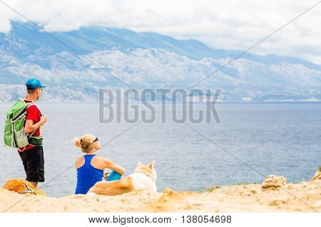 Happy couple hikers trekking with akita dog in summer mountains at seaside. Young woman and man walking on rocky mountain trail path looking at beautiful inspirational landscape view.