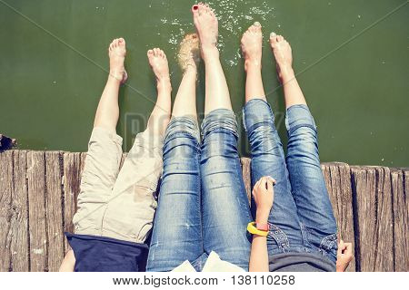 Two girls and boy have fun on pier splashing their legs into water of pond. Happy summer vacation background