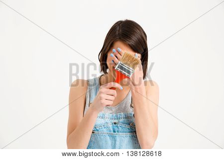 A portrait of young pretty girl, in gray shirt and denim overall, hiding face, holding painting brush, isolated on white background
