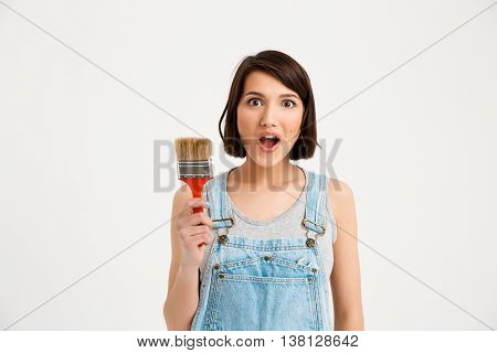 A portrait of young pretty girl, in gray shirt and denim overall, looking surprised, holding painting brush, isolated on white background