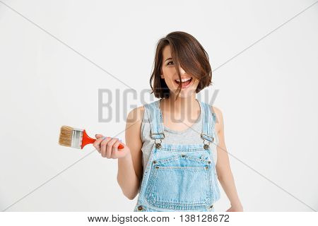 A portrait of young playful crazy beautiful girl, in gray shirt and denim overall, holding painting brush looking at camera, isolated on white background