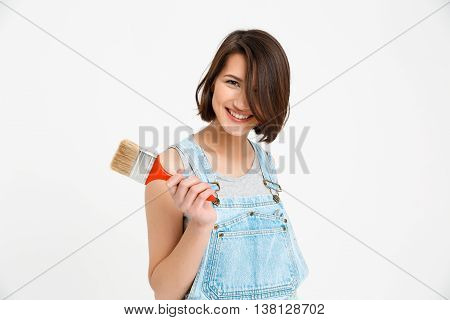 A portrait of young smiling  beautiful girl, in gray shirt and denim overall, holding painting brush looking at camera, isolated on white background