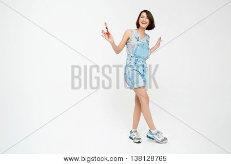 Full length portrait of a laughing pretty girl, in gray shirt and denim overall, holding painting brush, isolated on white background