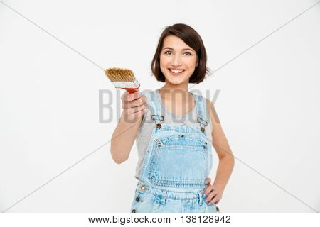 A portrait of young  beautiful girl, in gray shirt and denim overall, pointing forward with focus painting brush, looking at camera, isolated on white background