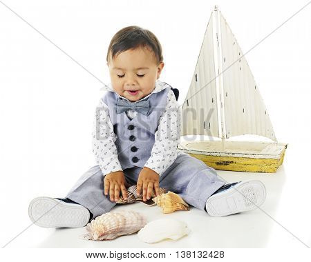 An adorable baby boy happily looking down at the sea shells he's playing with.  A toy sailboat is nearby.  On a white background.
