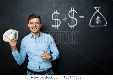 handsome man standing over blackboard. Holding dollars in his hands and smiling. thumbs up