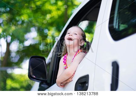 Little girl with funny pigtails watching out of car window sitting on front driver seat during a break on a family vacation road trip on summer day. Child in white minivan. Traveling by car with kids.