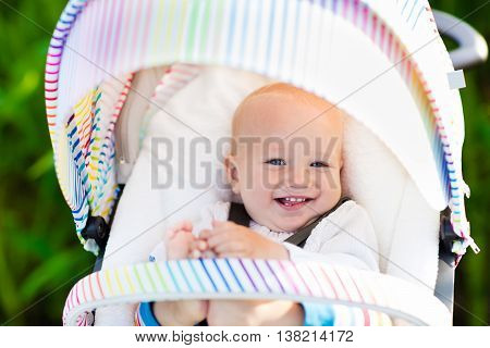 Baby boy in white sweater sitting in white stroller on a walk in a park. Child in colorful rainbow buggy. Little kid in a pushchair. Traveling with young kids. Transportation for family with infant.