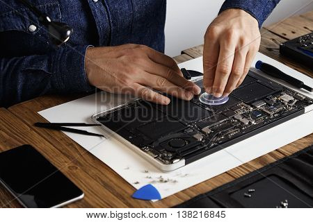Master uses small suction cup to change battery cells from broken laptop to repair and clean it in his laboratory with specific toolkit on wooden table around close focus on hands.