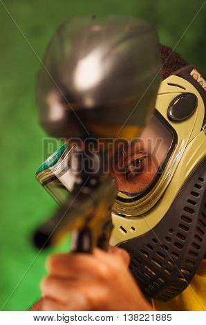Closeup headshot man wearing green and black protection facial mask facing camera pointing paintball gun.