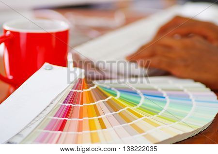 Closeup large palette colormap spread out in front of white computer keyboard and red coffee mug sitting on wooden desk, designer concept.
