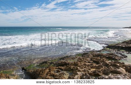Indian Ocean waters rushing the rocky beach under a blue sky with clouds at Penguin Island in Rockingham, Western Australia.
