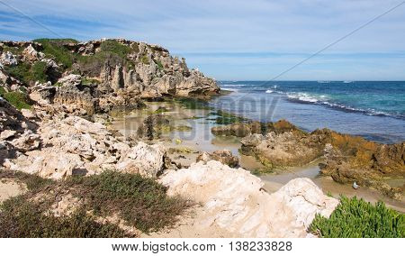 Indian Ocean seascape with rugged limestone bluffs and native flora under a blue sky with clouds at Penguin Island in Rockingham, Western Australia.