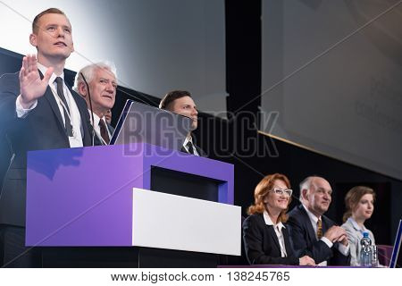 Young charismatic man speaking from a lectern during a conference surrounded by other participants