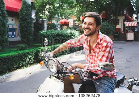 Handsome man riding a scooter on city roads. Summer potrait of male hipster with beard wearing a red shirt sitting on white retro motorbike