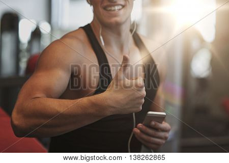 sport, bodybuilding, lifestyle, technology and people concept - happy man with smartphone and earphones listening to music and showing thumbs up in gym