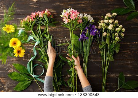 Bright colorful alstroemerias in hands, irises and roses on wood table, herberas in vase. From above.