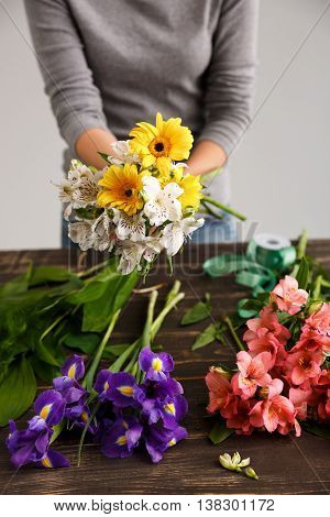 Girl in gray blouse and jeans make bouquet of herberas and alstroemerias over gray background, flowers on wood table.