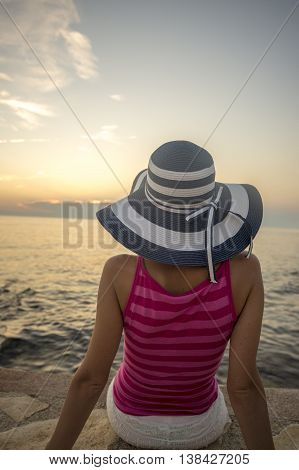 Rear view on person in pink sleeveless top and wide brim hat sitting on rock near water in front of setting or rising sun.