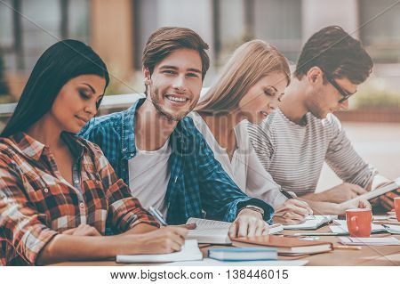 Preparing to exams together. Joyful young man smiling and looking at camera while sitting with his friends at the wooden desk outdoors