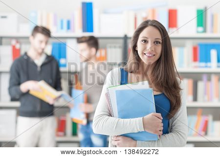 Confident smiling student girl posing in the library holding notebooks and looking at camera learning and education concept