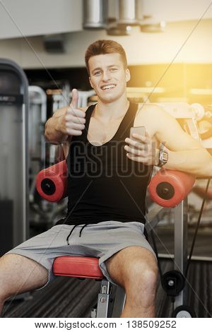 sport, bodybuilding, lifestyle, technology and people concept - smiling young man with smartphone showing thumbs up gesture in gym