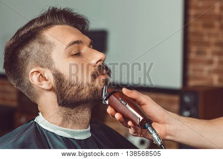 Hipster client visiting barber shop. The hands of young barber making the cut of beard