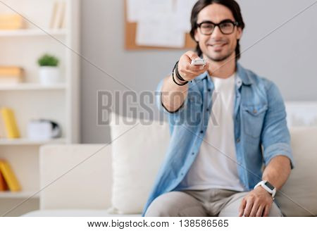 Switch it on. Pleasant handsome positive man sitting on the couch and holding remote control while watching TV
